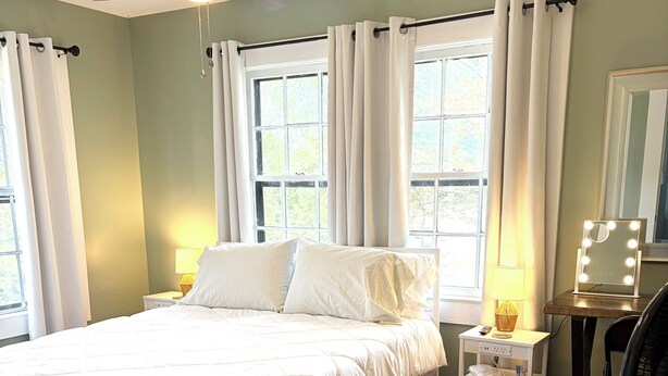 Ein Schlafzimmer mit Bett, Deckenventilator, Nachttisch mit Lampe, Spiegel mit Beleuchtung und Fenster mit Vorhängen.