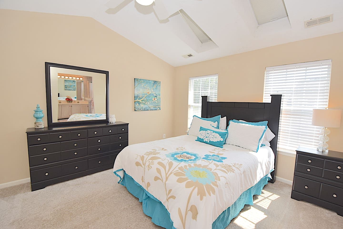 A bedroom with a bed, dresser, mirror, artwork, and windows.