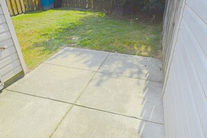 Eine betonierte Terrasse führt zu einem Haus mit Holzzaun und einem blauen Fass im Hintergrund.