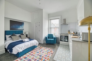 A bedroom with a bed, a blue armchair, a kitchen area with a microwave, and a window with blinds. - Glendale House by Guestz (Lytham-St-Annes)