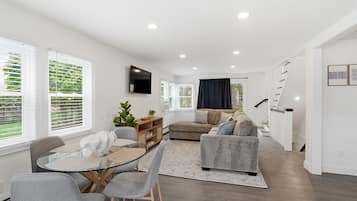 A modern living room with a glass dining table, grey sofas, and a staircase leading to another level.