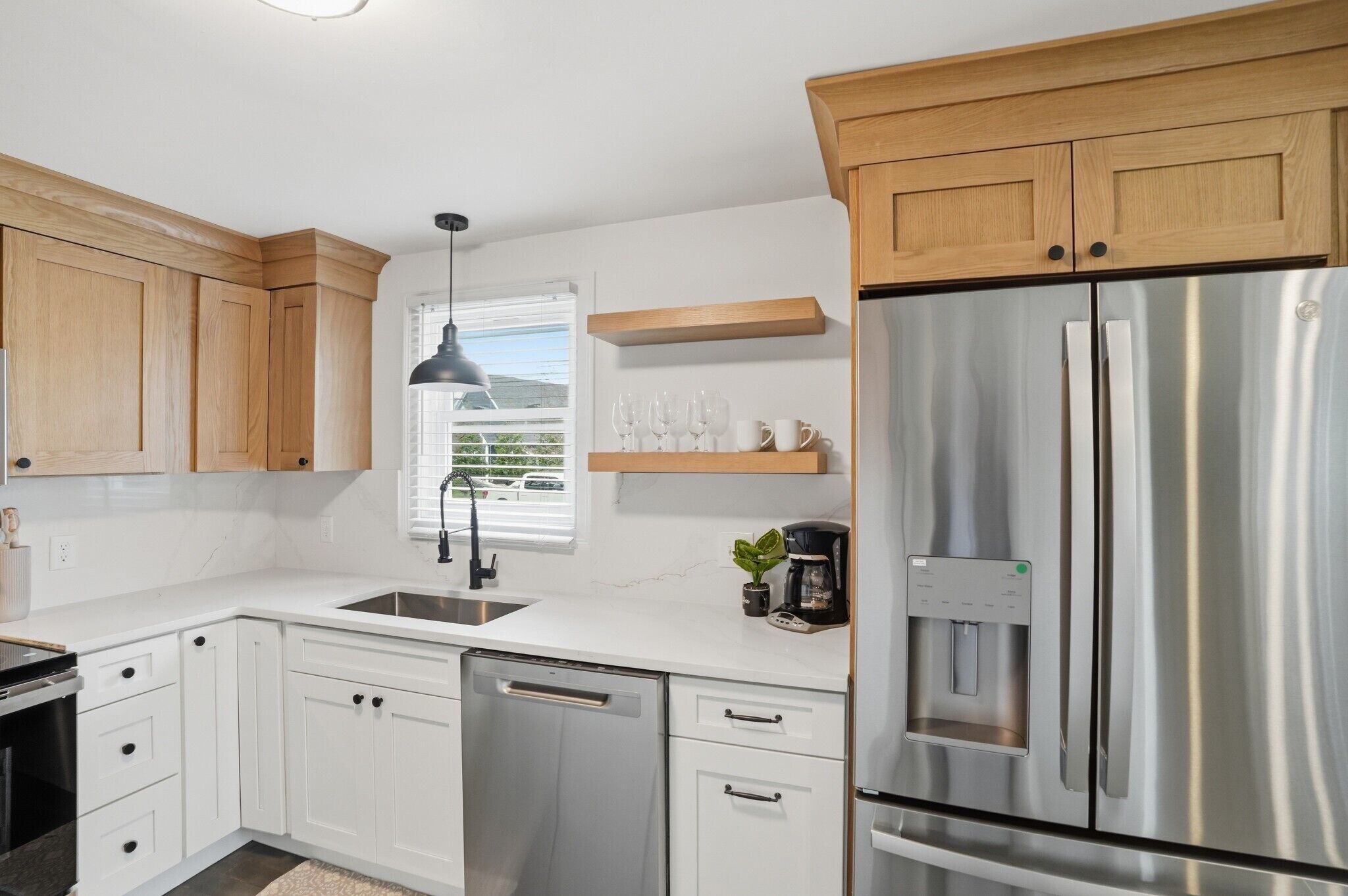 A modern kitchen with white cabinets, a stainless steel refrigerator, a built-in dishwasher, and a window with blinds.