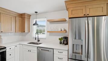 A modern kitchen with white cabinets, a stainless steel refrigerator, a built-in dishwasher, and a window with blinds.