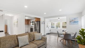 A modern living room with a sofa, dining table, and chairs, a kitchen in the background, and a large window with blinds.