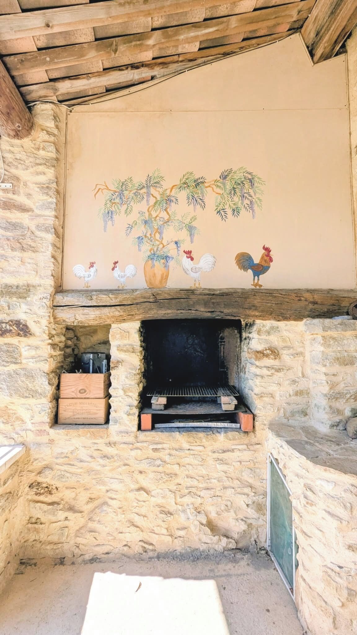 A stone fireplace with a mural of roosters and a tree, and a wooden chest labeled 'CHATEAU MAUCHARD.'
