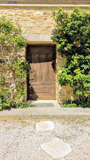 Ein rustikales Steinhaus mit einer Holztür, umgeben von viel Grün und einem Kiesweg.