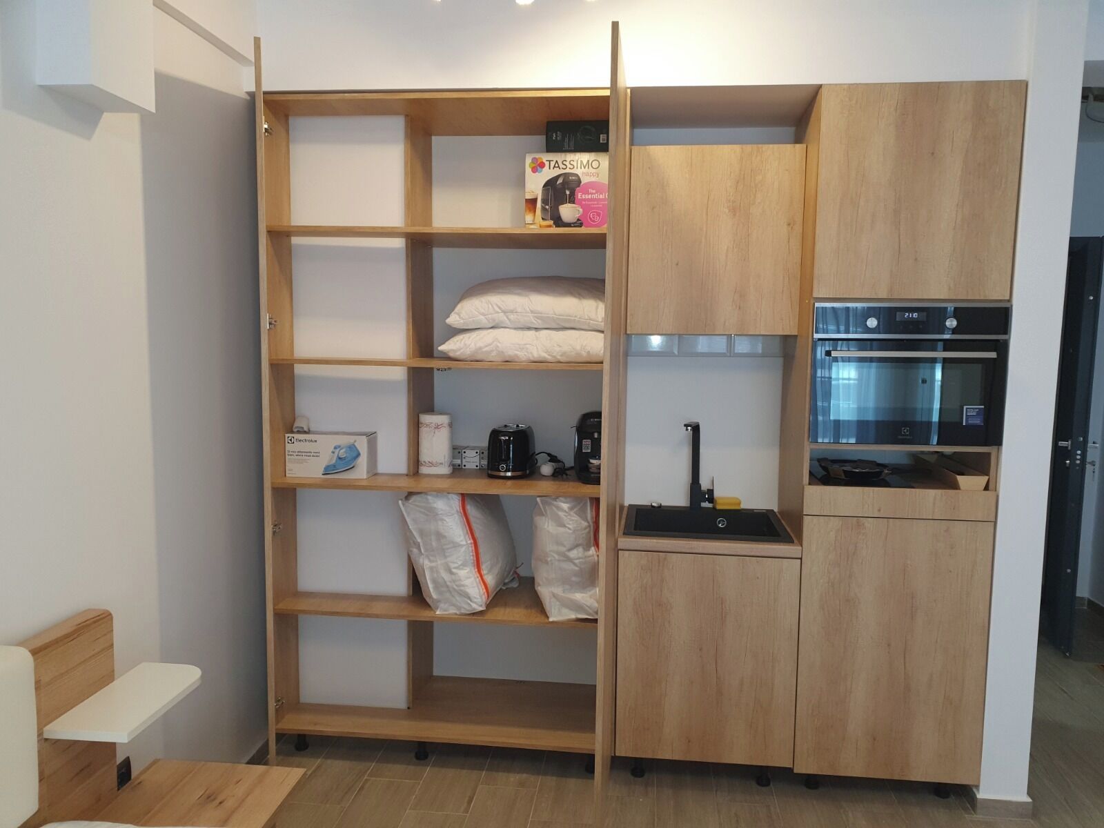 Een functionele keuken met een inbouwoven en magnetron, een houten plank met opbergzakken, en een klein aanrecht met waterkoker.