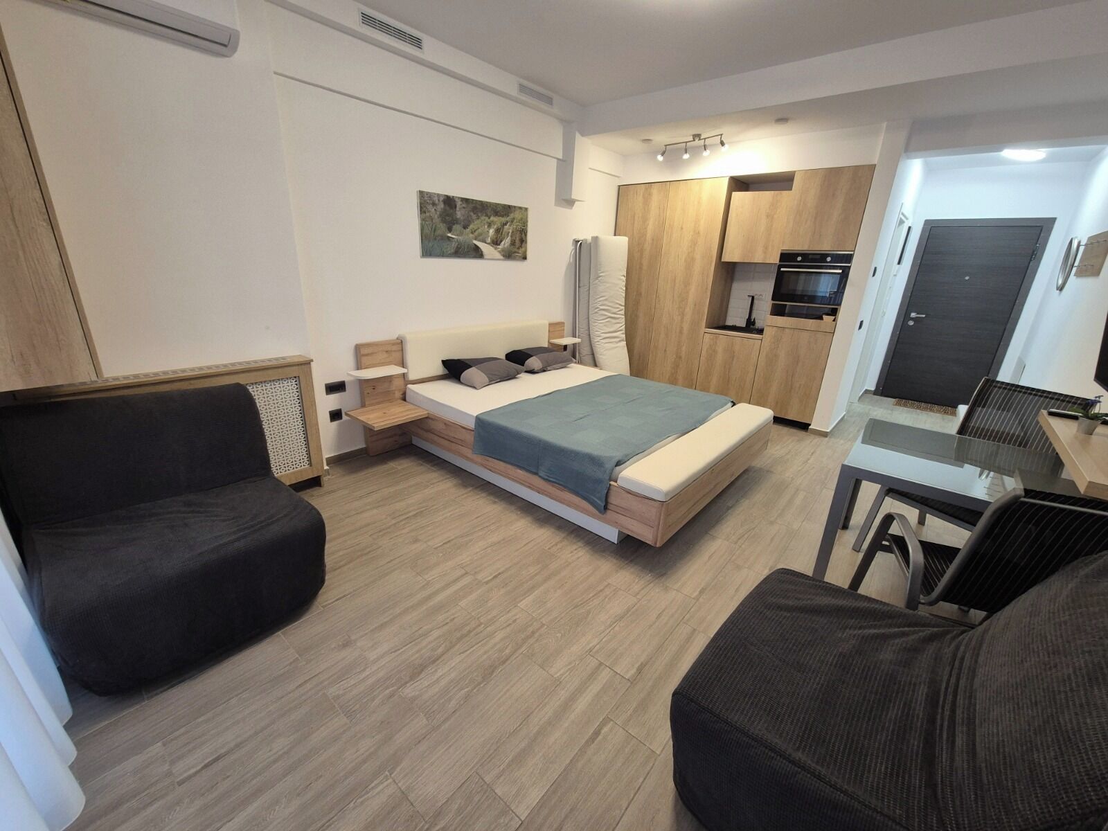 A modern bedroom with a bed, a small desk, a sofa, and a kitchenette.