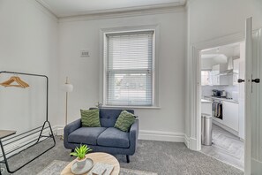 A living room with a grey sofa, a small round table with a plant, a hanging clothes rack, and a window with blinds. - Glendale House by Guestz (Lytham-St-Annes)
