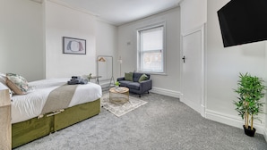 Une chambre à coucher avec un lit, un canapé, une petite table, une plante en pot et une fenêtre avec des stores.