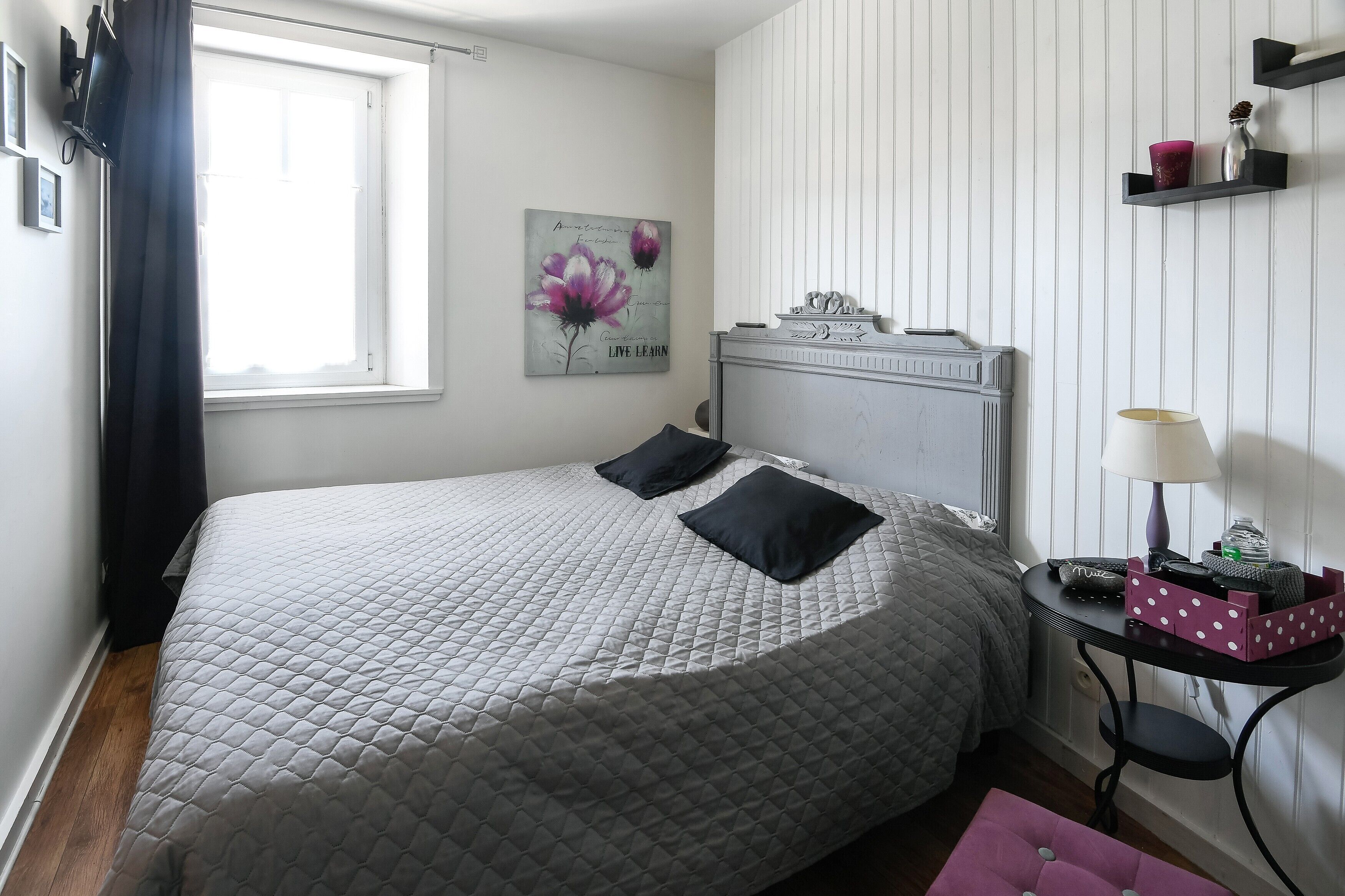 Ein Schlafzimmer mit Bett, Nachttisch, Lampe, Wandregal und einem Fenster mit Vorhängen.