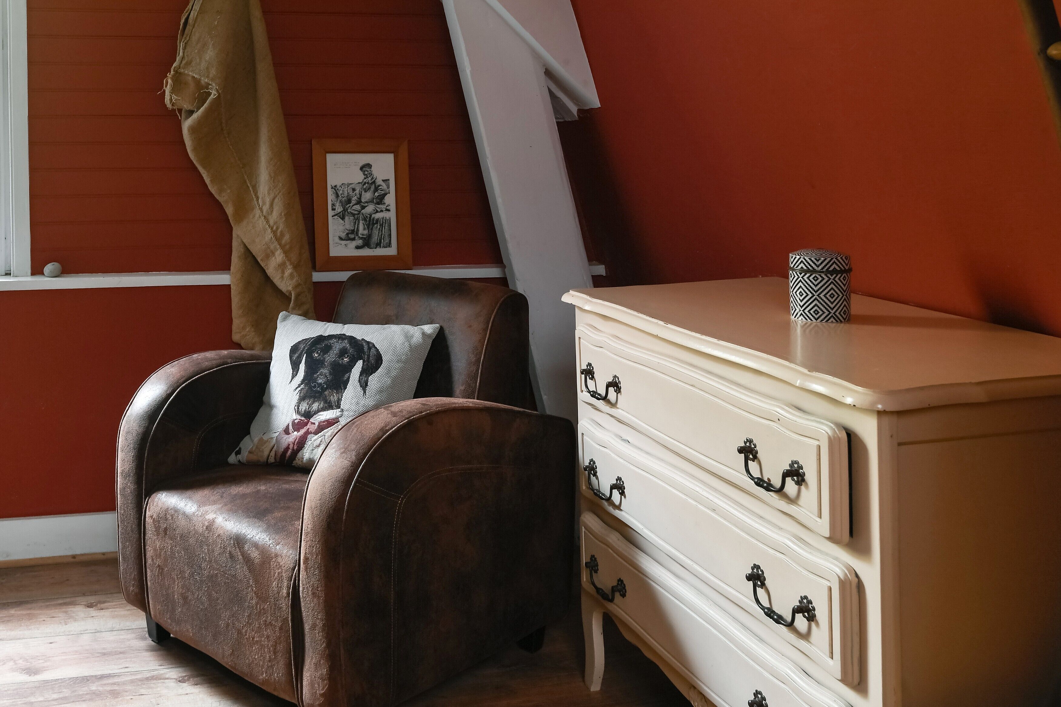 A room with a brown leather armchair, a white dresser with black handles, a framed picture on the wall, and a window with a curtain.