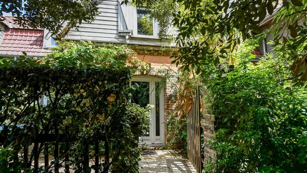Un étroit sentier mène à une maison dont la façade est en briques et la porte est peinte en blanc, le tout étant entouré d’une végétation luxuriante.