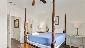A four-poster bed with a blue bedspread, a wooden bench, a desk with a lamp, and a ceiling fan. - Shell Seeker - Beachside Townhome - Just Steps from Beach (Mexico Beach)
