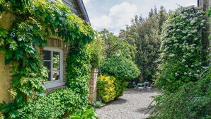 A narrow gravel path leading to a building with a window and a brick wall, surrounded by lush greenery and white flowering plants. - Cottages de la Croix l'Abbé, la Bergeronnette – Tranquil Duplex Retreat (Saint-Valery-sur-Somme)