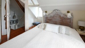 A bedroom with a large bed, a wooden headboard, a nightstand with a lamp, and a view of a staircase. - Cottages de la Croix l'Abbé, la Bergeronnette – Tranquil Duplex Retreat (Saint-Valery-sur-Somme)