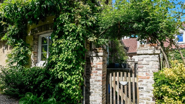 A stone gate with a wooden fence, surrounded by lush greenery and a building with a window. - Cottages de la Croix l'Abbé, la Bergeronnette – Tranquil Duplex Retreat (Saint-Valery-sur-Somme)