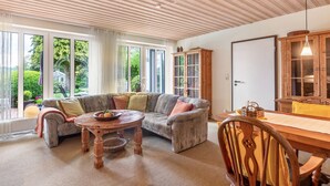 Ein Wohnzimmer mit Holzboden, großen Fenstern, einer Couch, einem Esstisch und einem Schrank.