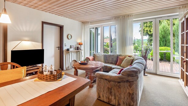 Ein Wohnzimmer mit Holzboden, einem Fernseher, einem Esstisch mit Korb, einer Couch und Blick ins Freie durch große Fenster.