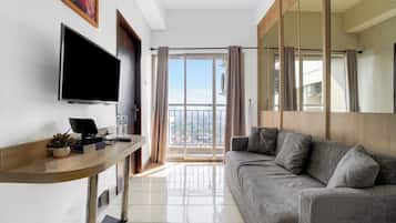 A modern living room with a flat-screen TV, a sofa, a small table with a keyboard and a plant, and a sliding glass door leading to a balcony.
