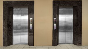 Dos puertas de ascensor con pantallas digitales, frente a una pared lisa con suelo de baldosas.