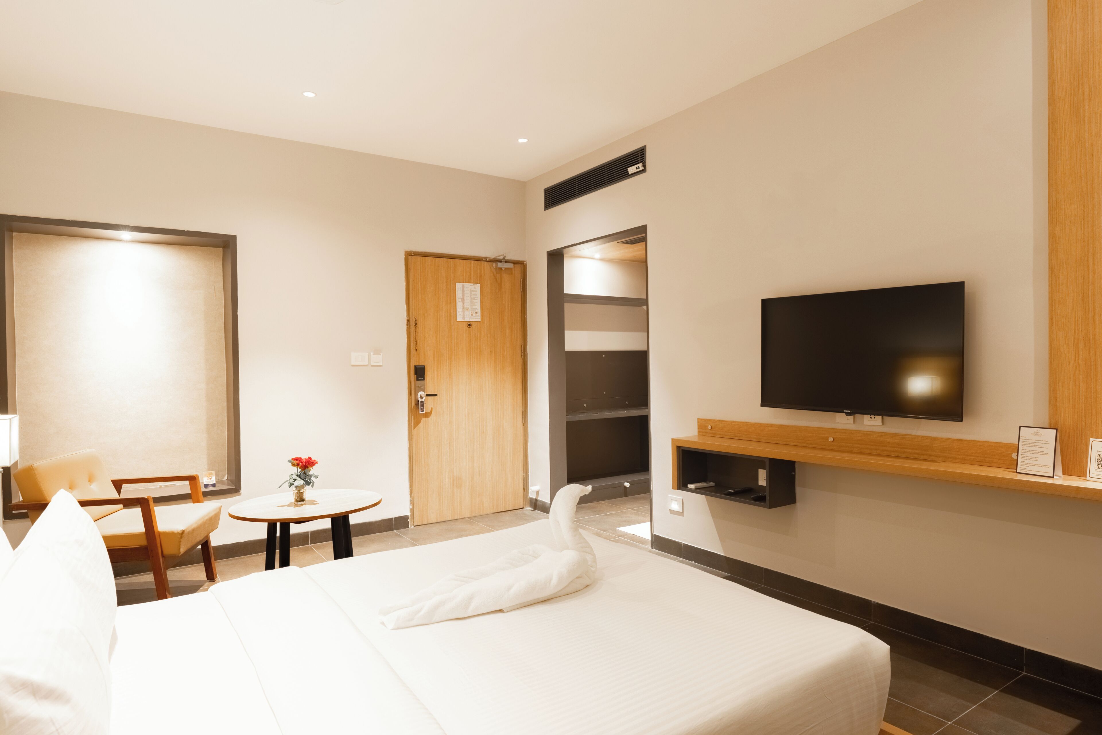 Habitación de hotel con cama, televisión, puerta de madera, una pequeña mesa con un jarrón y una silla.