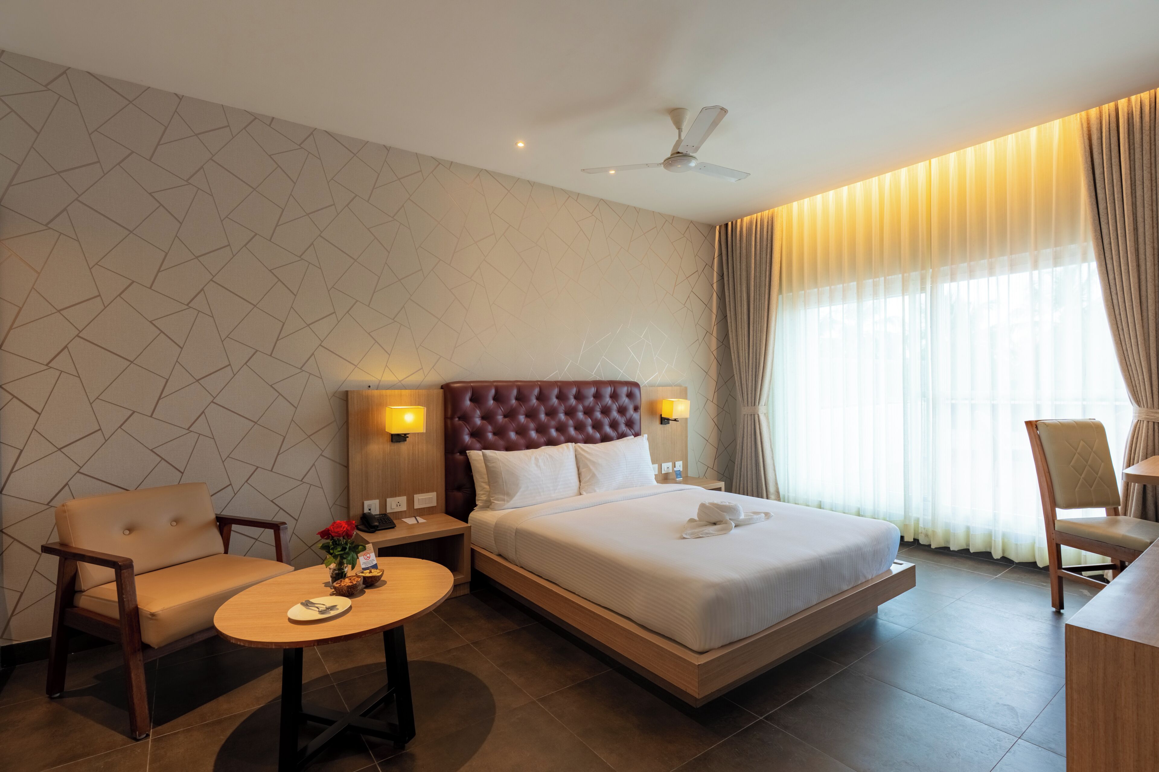 A hotel room with a bed, a chair, a small table, a ceiling fan, and a window with curtains.