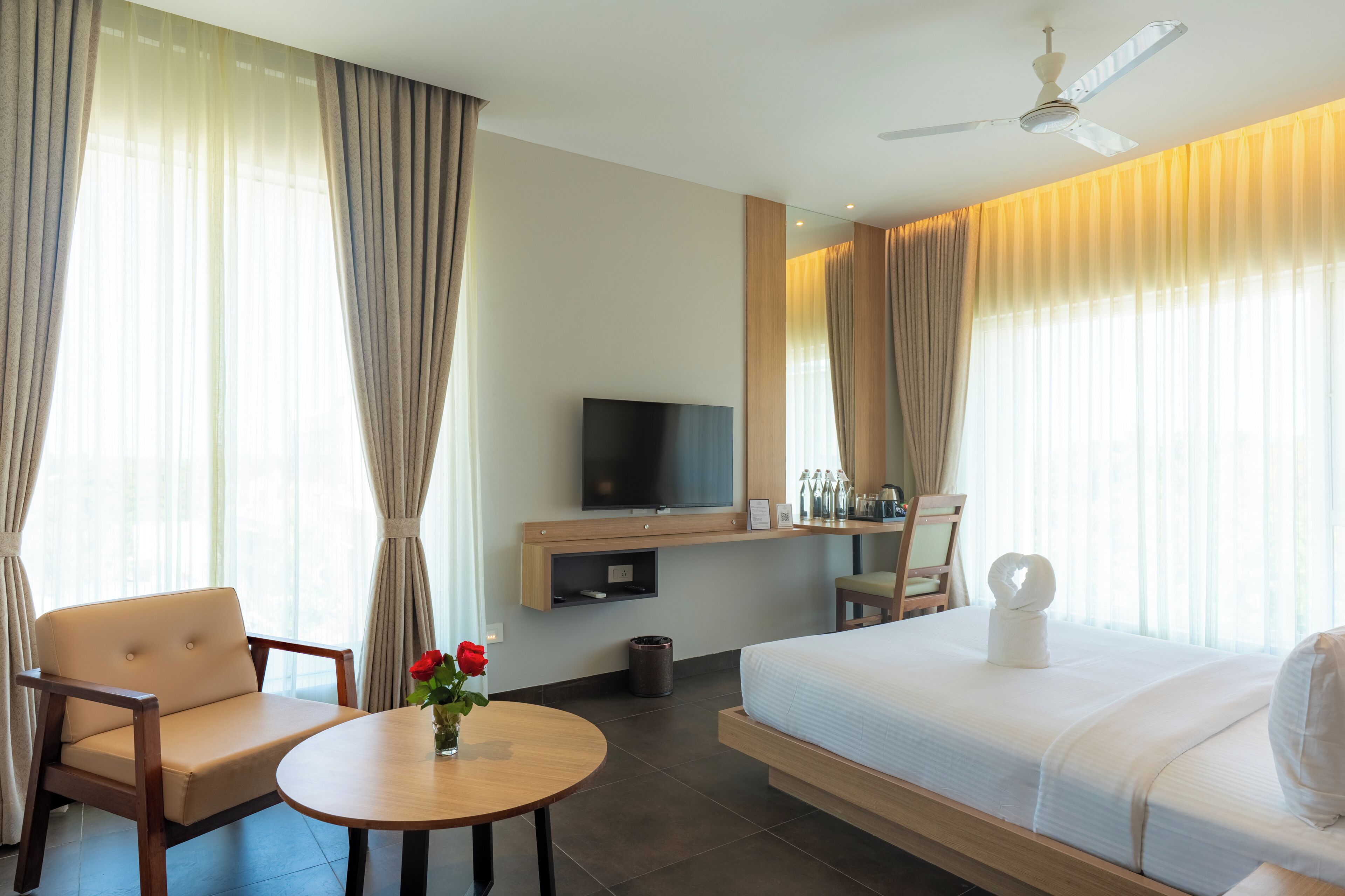 A hotel room with a bed, a TV, a desk, a chair, a table, and a vase with flowers.