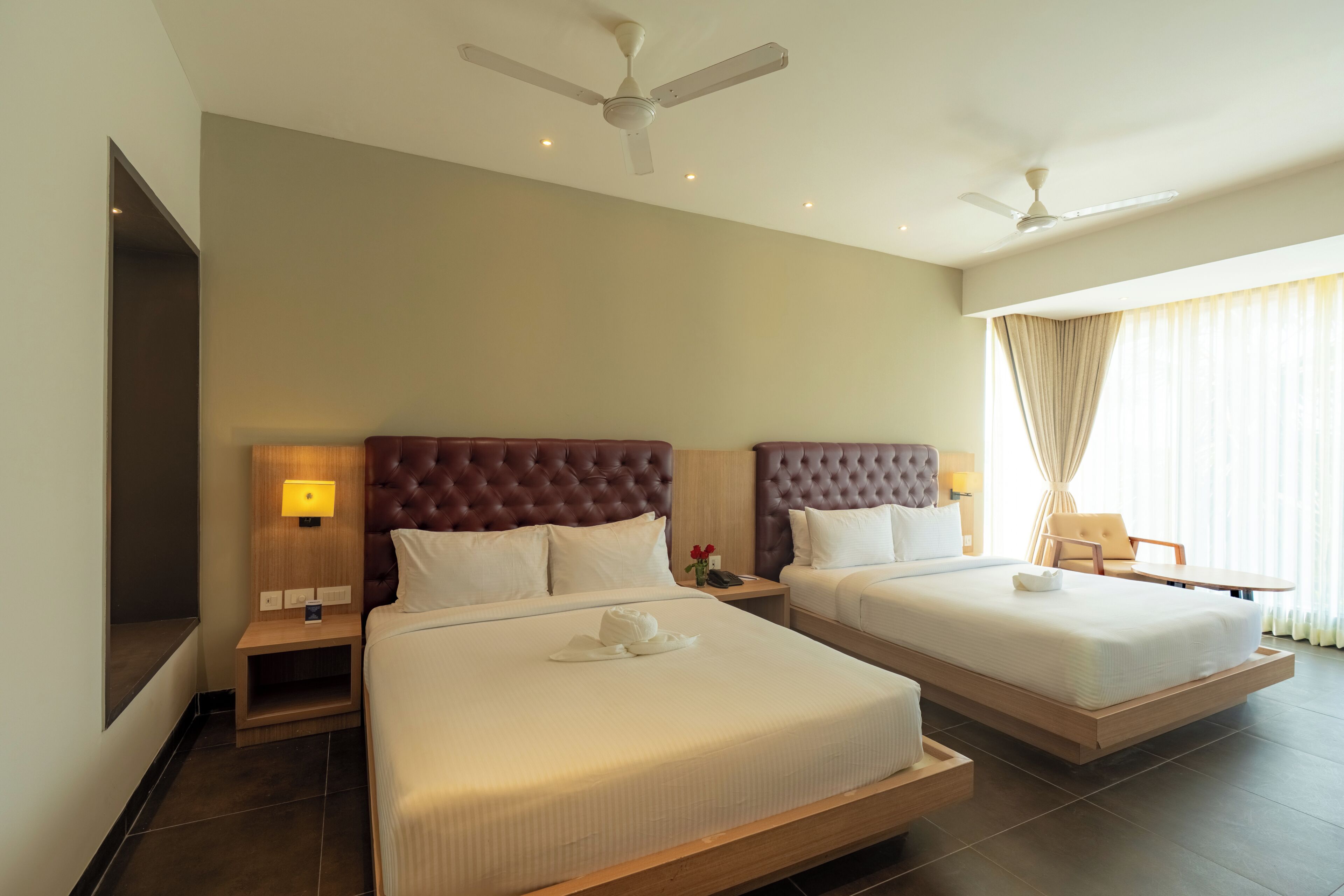 Habitación de hotel con dos camas, ventilador de techo, televisor y ventana con cortinas.