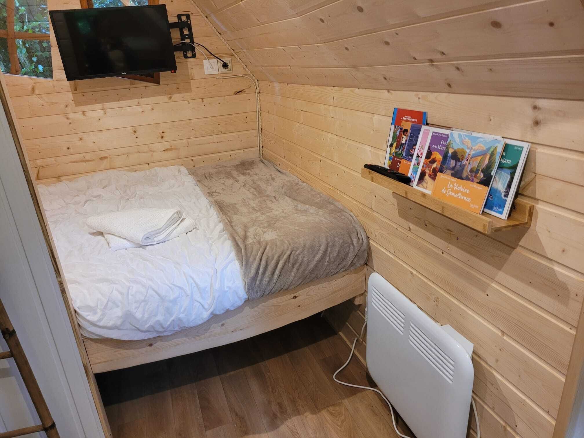 Una piccola stanza in legno con un letto, una TV a parete, un termosifone e uno scaffale con libri.