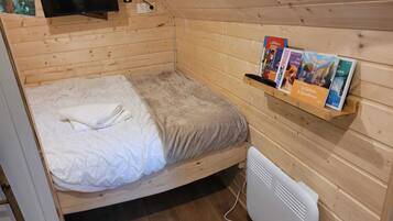 Una habitación pequeña de madera con una cama, un televisor montado en la pared, un calentador y una estantería con libros.