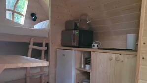 A compact kitchen with wooden walls, a refrigerator, a microwave, a kettle, and a small table with benches.