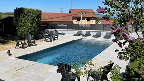 A swimming pool with lounge chairs, a hedge, and a building with a red roof in the background.