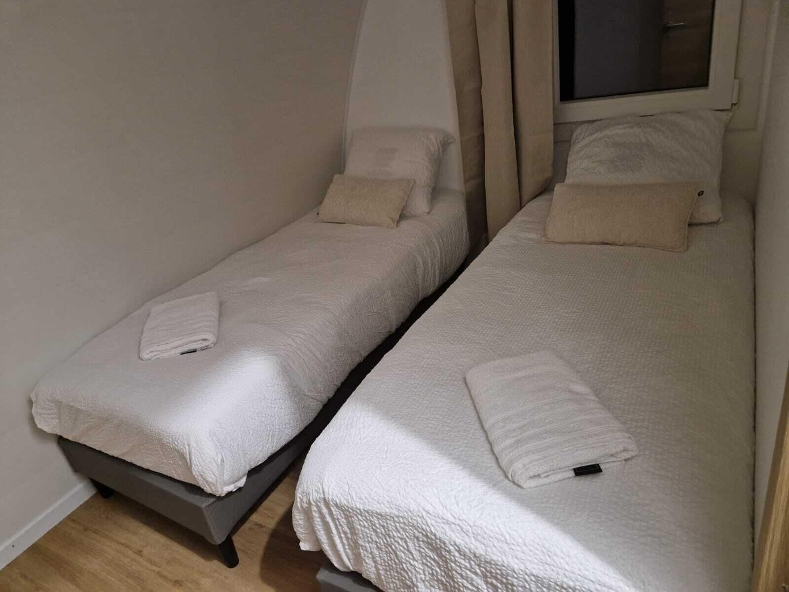 Dos camas individuales con sábanas y almohadas blancas, una ventana con cortinas y suelo de madera.