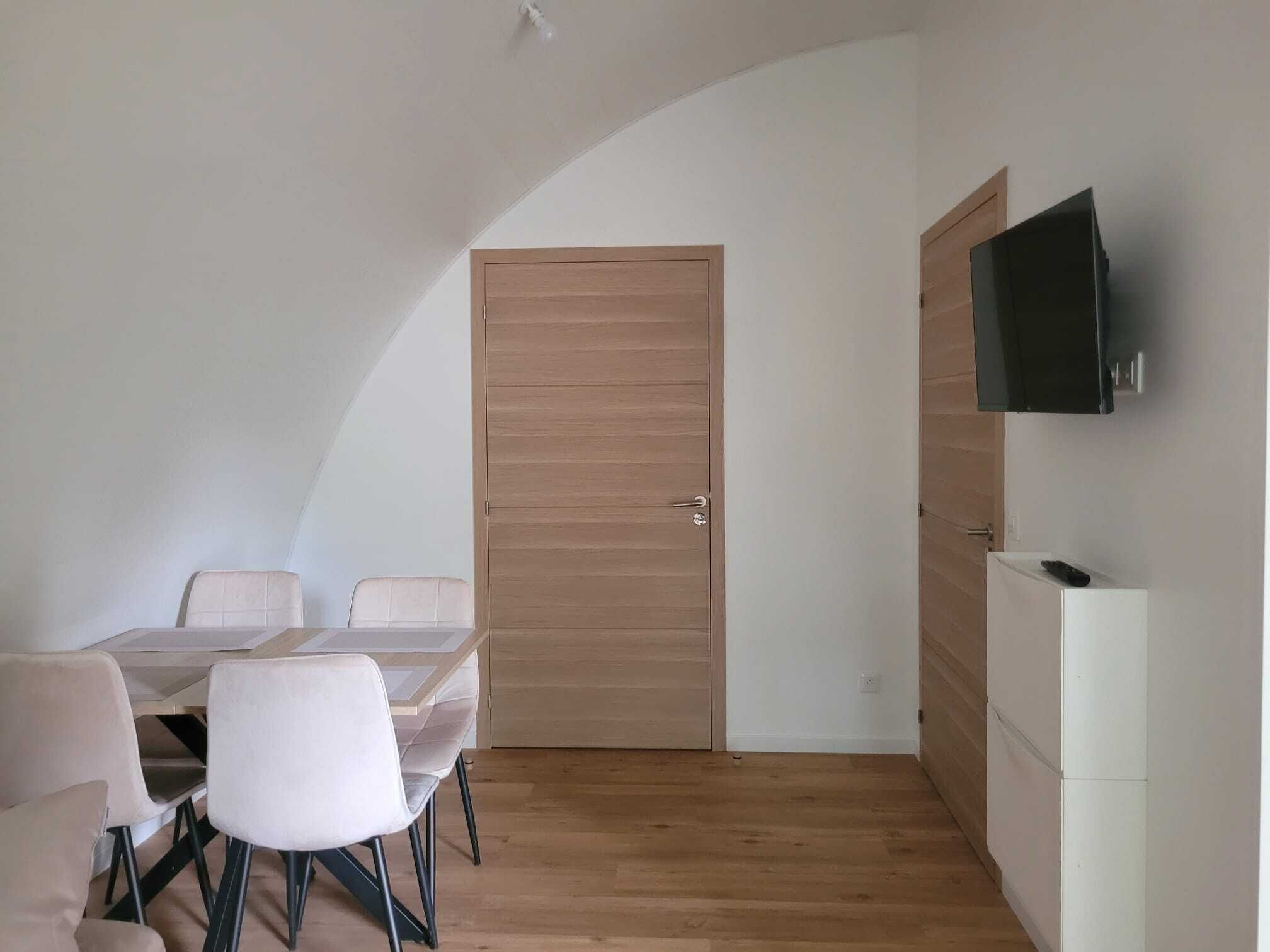 A room with a dining table, chairs, a wooden door, a white cabinet, and a mounted television.
