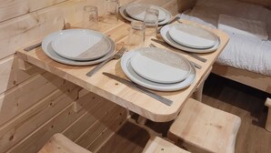 A wooden interior with a dining table set for four, benches, and a sleeping area with a bed.