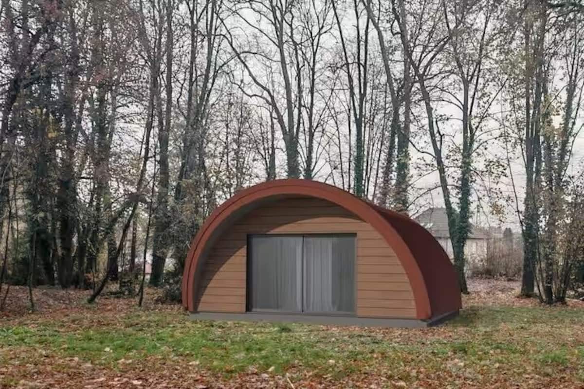Una pequeña estructura de madera semicircular con una puerta gris, rodeada de árboles.