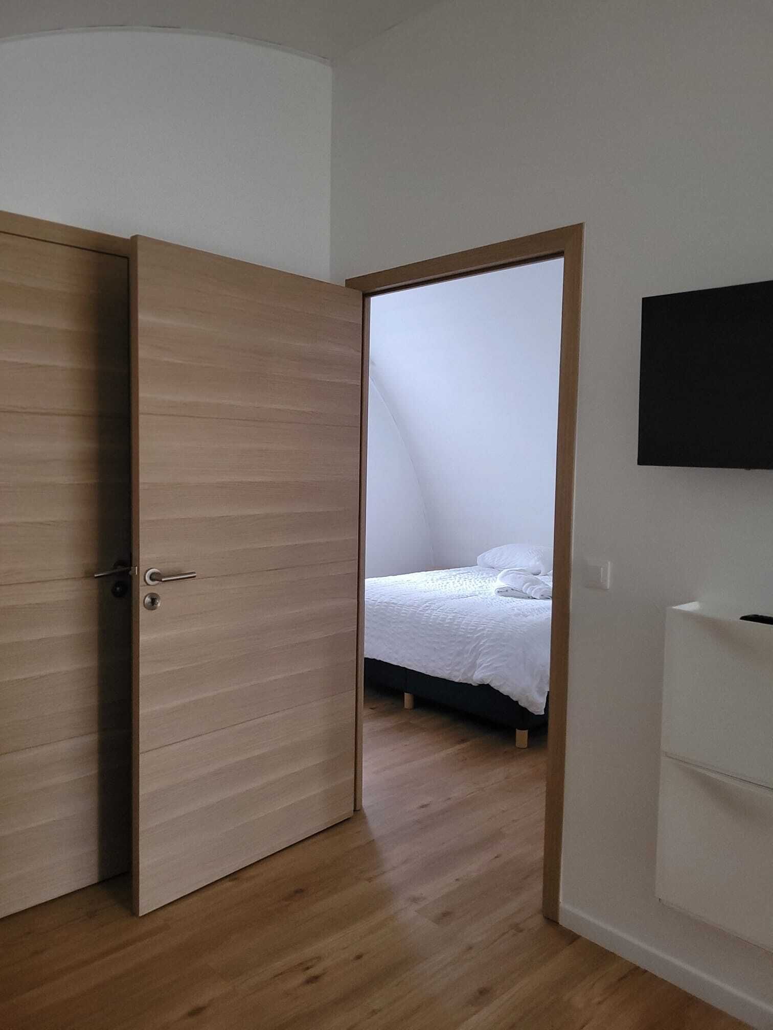A room with a wooden floor, a bed with white bedding, a closed door, and a flat-screen TV mounted on the wall.