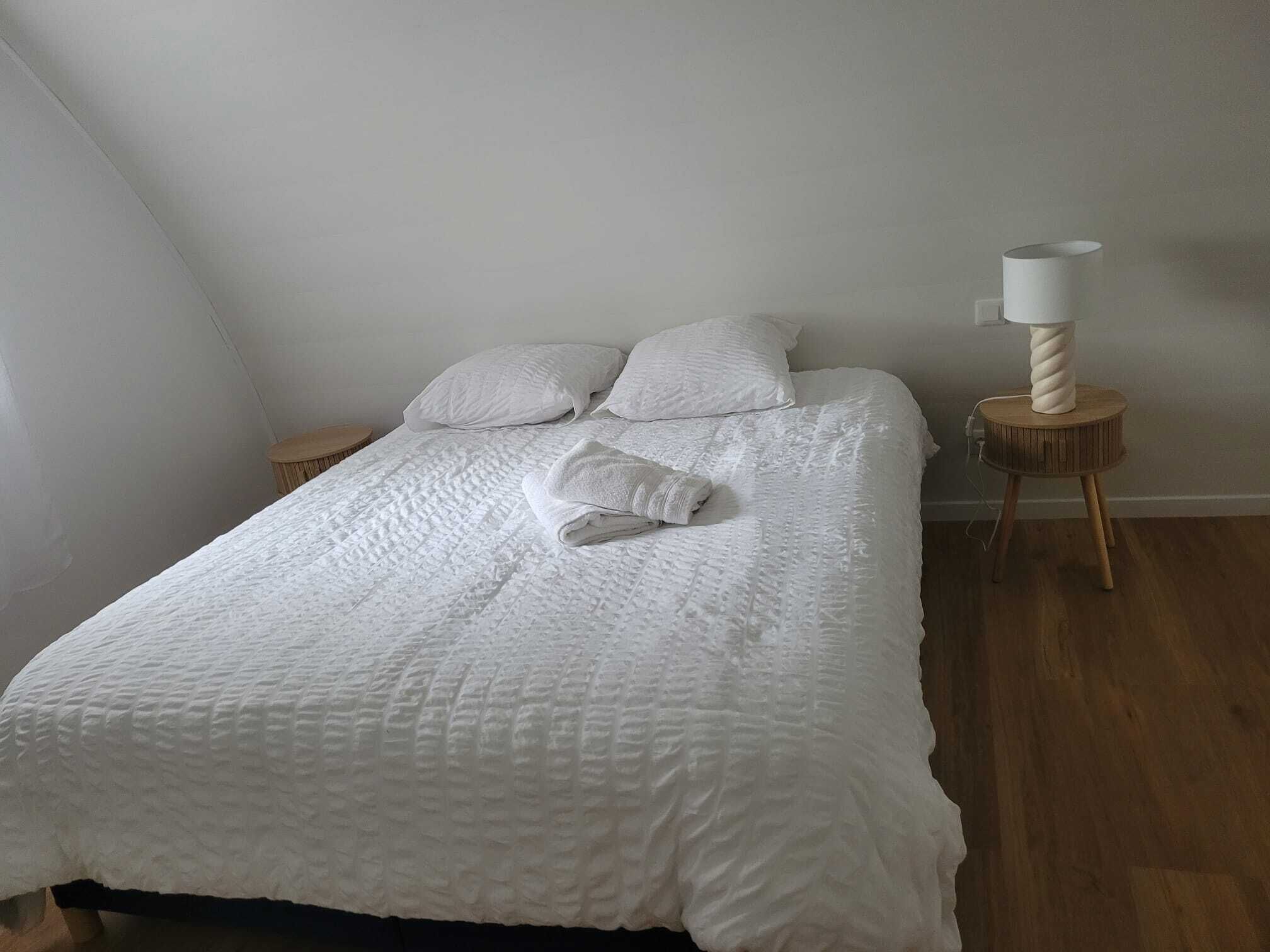 Ein ordentlich bezogenes Bett mit weißer Bettdecke und Kissen, ein gefaltetes Handtuch, eine Nachttischlampe und ein runder Nachttisch aus Holz.
