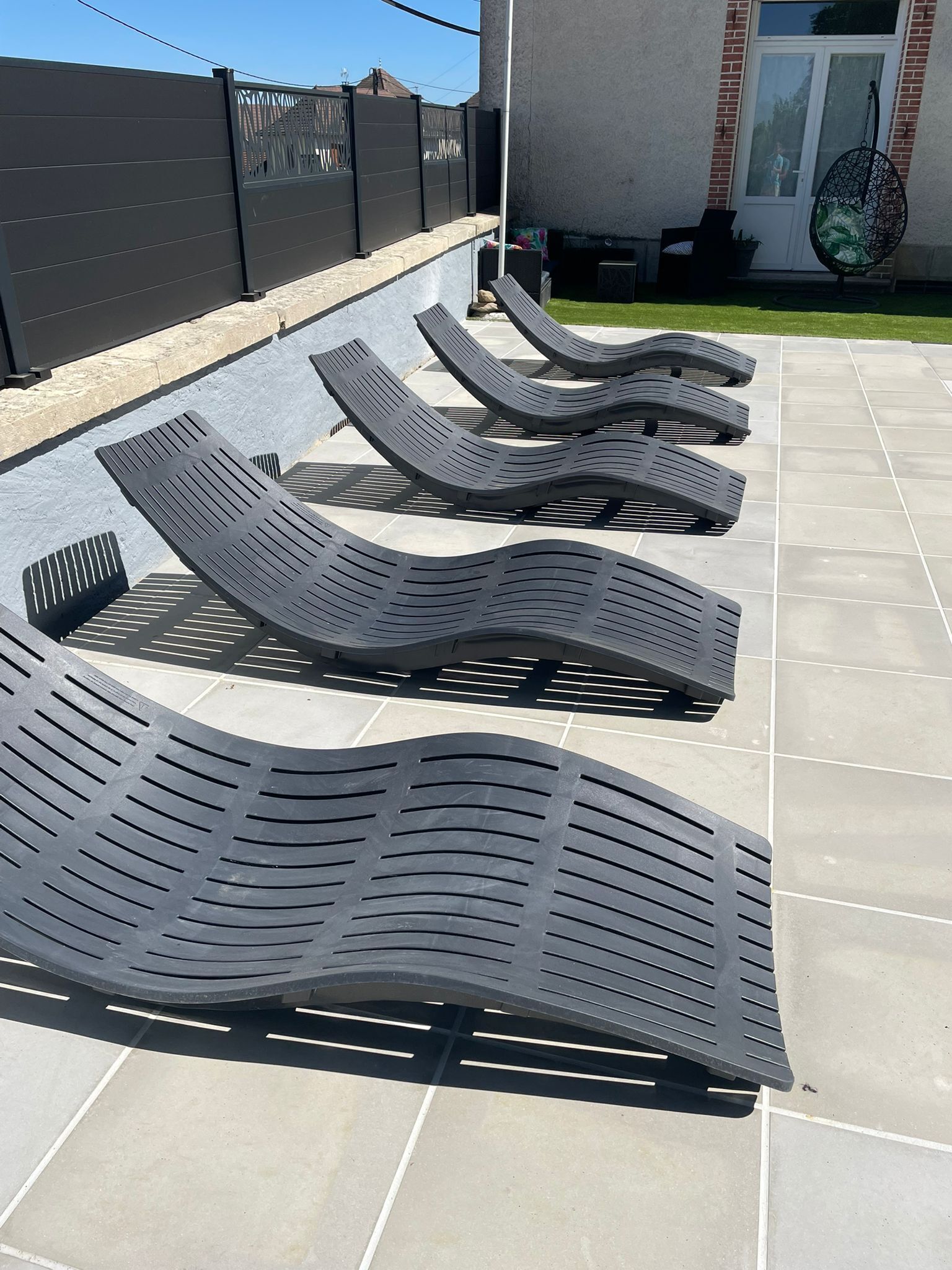 Outdoor lounge chairs arranged on a patio with a tiled floor, a black fence, and a building with a glass door in the background.