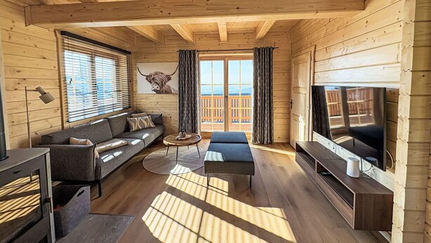 Ein Holzblockhaus-Interieur mit einer Couch, einem Fernseher und einem großen Fenster mit Blick auf eine Terrasse.
