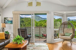 Ein Zimmer mit Blick auf üppiges Grün und Berge, eine große Schiebeglasdoor, ein Ledersofa, ein Holztisch und ein Korbstuhl.