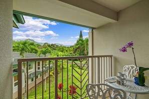 A balcony with a view of a lush garden and distant mountains, a white table with chairs, and a potted orchid. - Island Living at Villas of Kamalii 39 (Princeville)