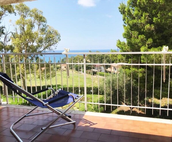 A balcony with a metal railing, a blue lounge chair, and a view of a green landscape and the sea.