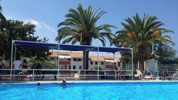 A swimming pool with a covered area, palm trees, and a building with a tiled roof in the background. - Residence La Giungla (Falerna)