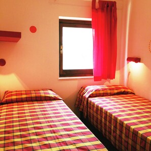 A room with two beds, a window with a curtain, and a wall-mounted light fixture.