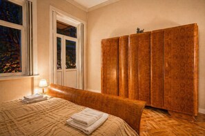 Quarto de hotel com cama, mesa de cabeceira, luminária, janela com vista para árvores e guarda-roupa.