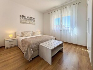 Ein Schlafzimmer mit Bett, Nachttischen, einer Bank, einem Fenster mit Vorhängen und einem Wandbild.