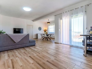 Ein modernes Wohnzimmer mit grauem Sofa, Holzboden, Flachbildfernseher, Essbereich mit Tisch und Stühlen sowie Balkon mit Geländer.