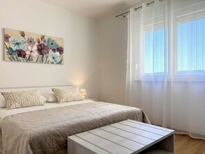 Ein Schlafzimmer mit Bett, Nachttisch, Lampe, Fenster mit durchscheinenden Vorhängen und einem floralen Bild an der Wand.